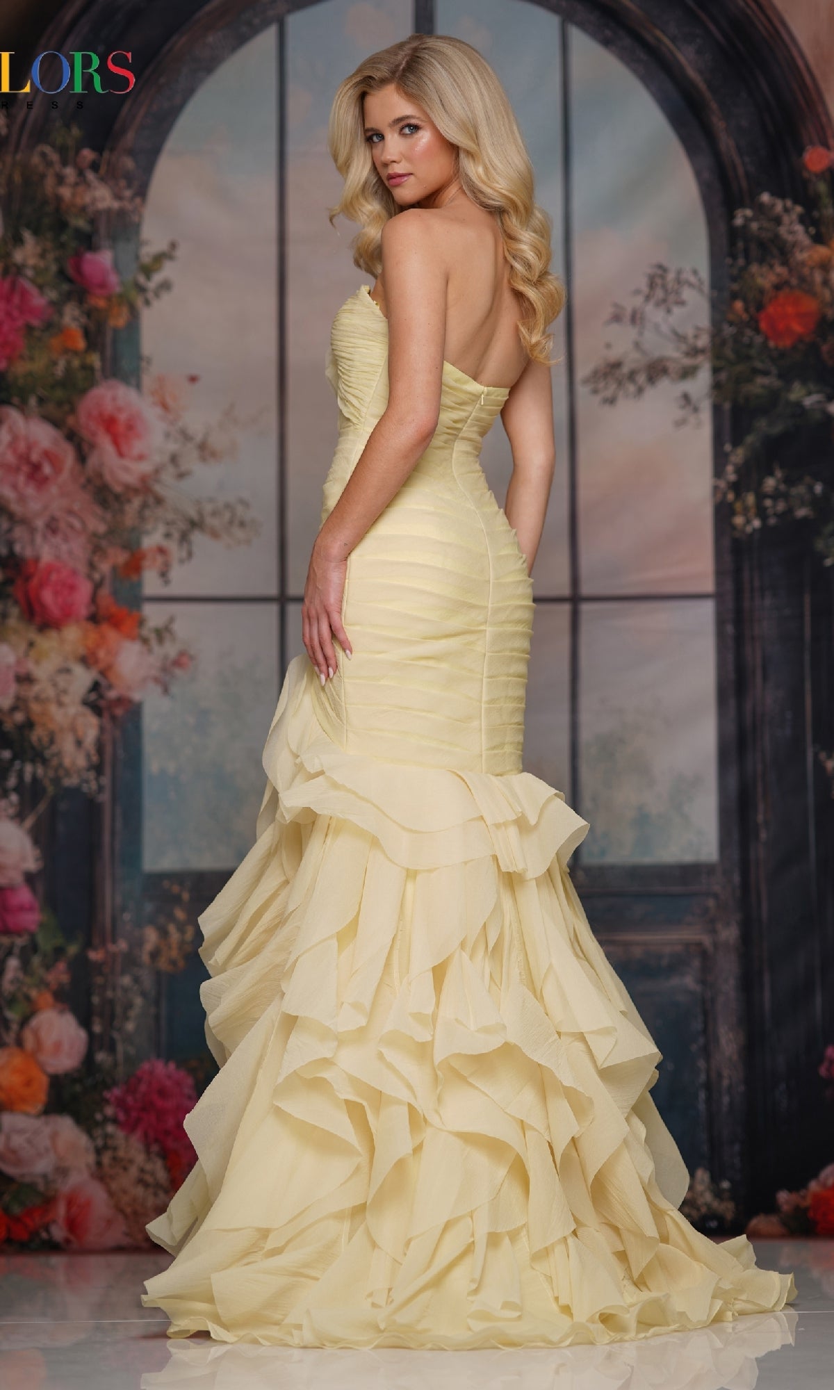 A woman wearing the Strapless Chiffon Prom Gown: Colors Dress 3886 poses before a floral, arched window backdrop, looking over her shoulder in a fitted yellow mermaid gown with a ruffled skirt.